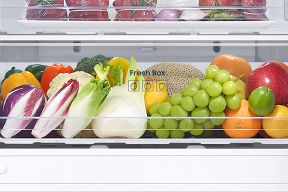 Szuflada Fresh Box Wnętrze lodówki z szufladą wypełnioną różnorodnymi warzywami i owocami, takimi jak winogrona, jabłka, pomarańcze, sałata i brokuły. Na szufladzie widoczny jest napis \'Fresh Box\' z ikonami.