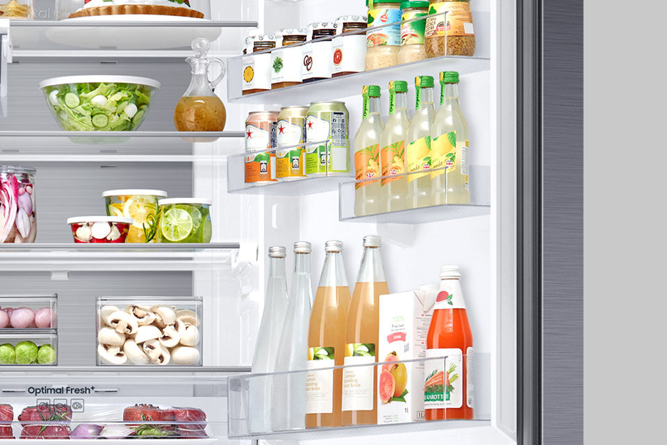Wnętrze lodówki z półkami wypełnionymi różnorodnymi produktami spożywczymi, takimi jak napoje w butelkach, słoiki z przetworami oraz świeże warzywa i owoce. Na drzwiach lodówki znajdują się dodatkowe półki z napojami i przyprawami.