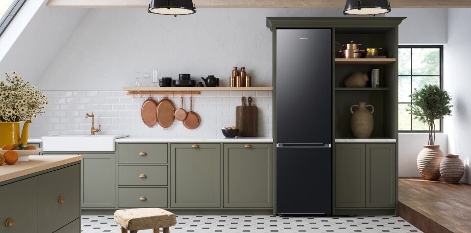 Kuchnia z zielonymi szafkami, białym zlewem i czarną lodówką marki Samsung. Na blacie i półkach znajdują się różne akcesoria kuchenne, a na podłodze jest biało-czarna mozaika.