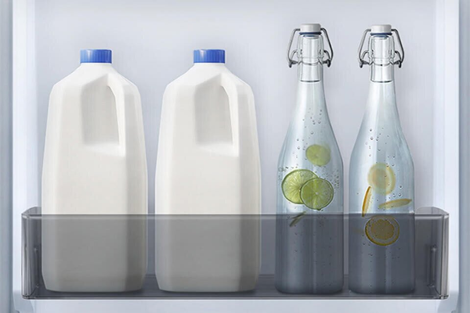 W lodówce znajdują się dwa kartony z mlekiem z niebieskimi zakrętkami oraz dwie szklane butelki z wodą i plasterkami cytryny i limonki. Butelki i kartony są ustawione w przezroczystej półce na drzwiach lodówki.