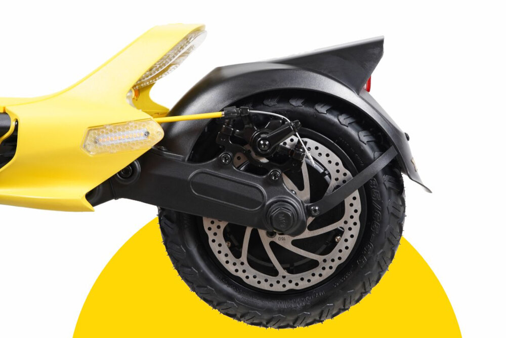 Zdjęcie przedstawia tylne koło żółtej hulajnogi elektrycznej z widocznym amortyzatorem i tarczą hamulcową. Tło jest białe z żółtym półokręgiem pod kołem.