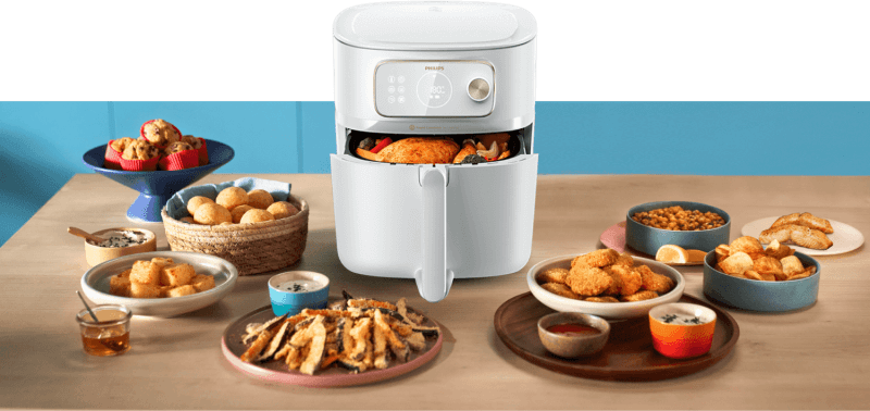 Na stole znajduje się frytkownica beztłuszczowa z otwartą szufladą, w której widać pieczone mięso. Wokół frytkownicy rozmieszczone są różne potrawy, takie jak pieczywo, frytki i muffiny.