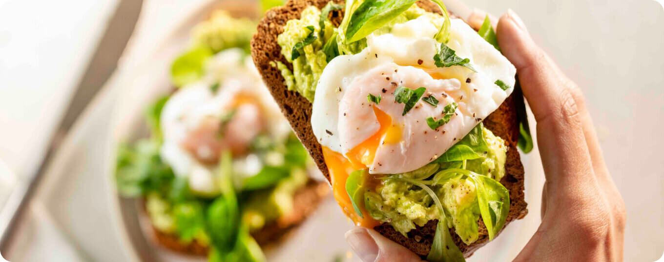 Osoba trzyma kanapkę z ciemnego pieczywa z awokado, jajkiem w koszulce i ziołami. W tle widoczna jest druga podobna kanapka na talerzu.