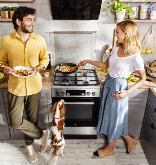 Kuchnia gazowa z czterema palnikami, na której gotowane jest jedzenie na patelni. W tle widoczne są szafki kuchenne i różne akcesoria kuchenne.