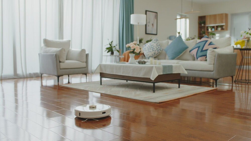 W przestronnym salonie z drewnianą podłogą znajduje się robot sprzątający. W tle widać sofę z poduszkami, stolik kawowy z obrusem i dekoracjami oraz zasłony przy oknach.