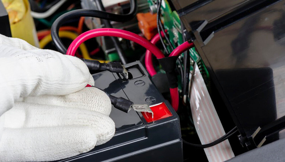 Osoba w białej rękawicy podłącza przewody do akumulatora, który jest częścią większego układu elektronicznego. Widoczne są czerwone i czarne przewody oraz elementy elektroniczne w tle.