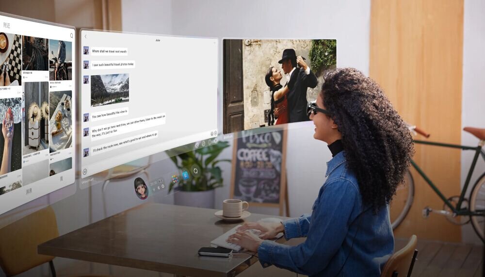 Osoba siedzi przy stole, nosząc okulary, przed nią wyświetlane są wirtualne ekrany z różnymi treściami, w tym zdjęciami i wiadomościami tekstowymi. W tle widać rower oparty o ścianę.