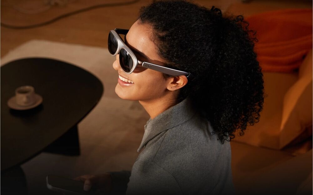 Osoba nosi czarne okulary ROKID Max 2, siedząc w pomieszczeniu z ciemnym stolikiem i filiżanką. W tle widoczna jest pomarańczowa poduszka na sofie.