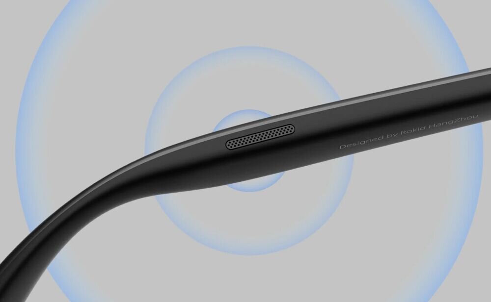 Czarna oprawka okularów z widocznym napisem \'Designed by Rokid Hangzhou\' oraz małym otworem wentylacyjnym. W tle widoczne są koncentryczne, niebieskie okręgi.