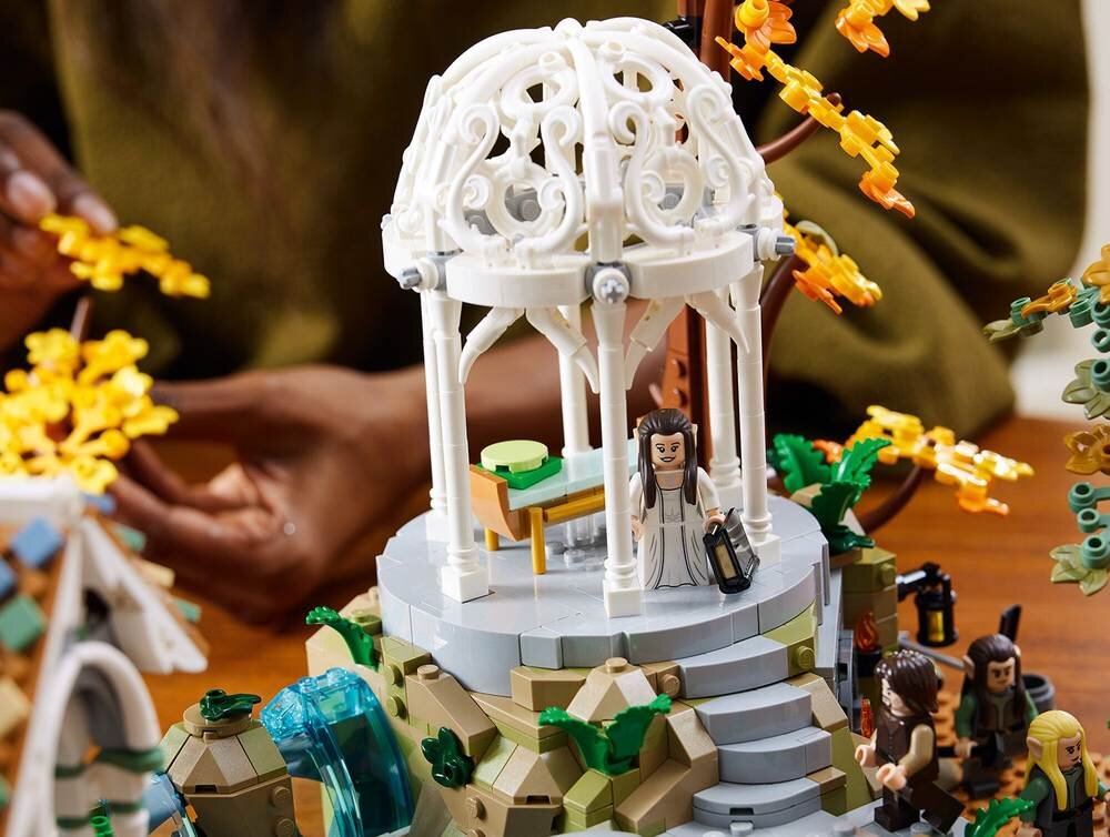 Zestaw LEGO przedstawiający scenę z altaną i figurkami, otoczony elementami przypominającymi roślinność i skały. W tle widoczne są ręce osoby układającej klocki.