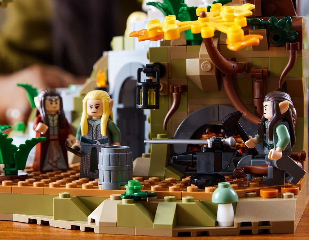 Na obrazie widoczne są figurki LEGO przedstawiające postacie fantasy w scenerii zbudowanej z klocków, z elementami roślinnymi i architektonicznymi. W tle znajduje się kowadło i miecz, a na pierwszym planie beczka i grzyb.