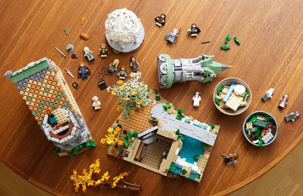 Na drewnianym stole znajdują się elementy zestawu LEGO, w tym miniaturowe figurki postaci oraz części budowli z motywem Rivendell. Widoczne są także miseczki z drobnymi klockami i akcesoriami.