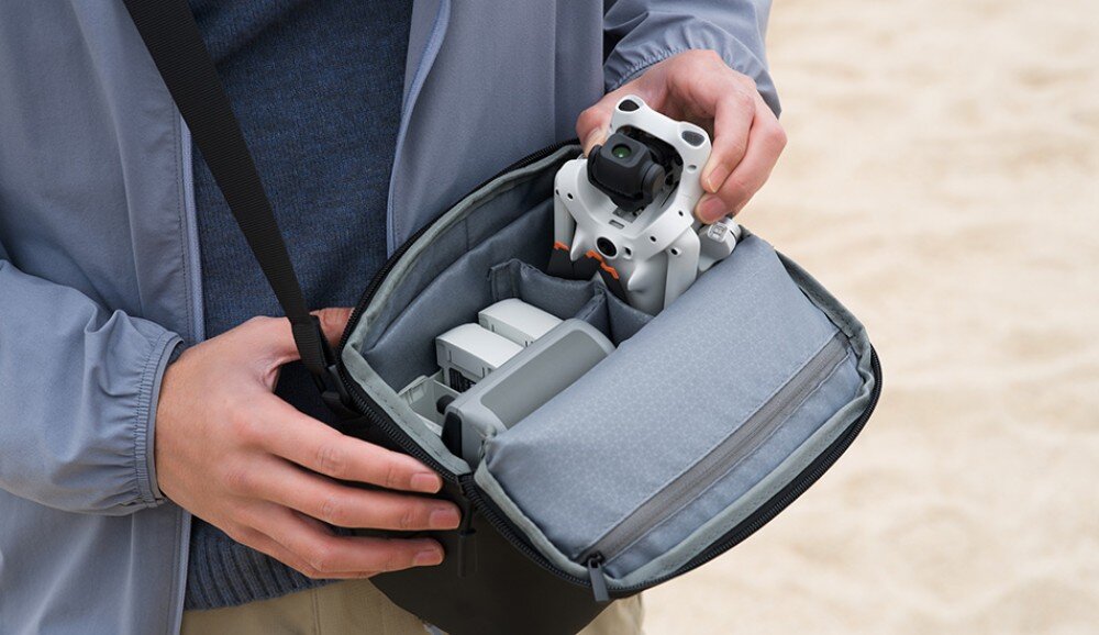 Dron DJI Lito 1 Osoba w szarej bluzie trzyma otwartą czarną torbę na ramię i wyciąga z niej szarego drona z czterema złożonymi śmigłami. W torbie widać również białą baterię