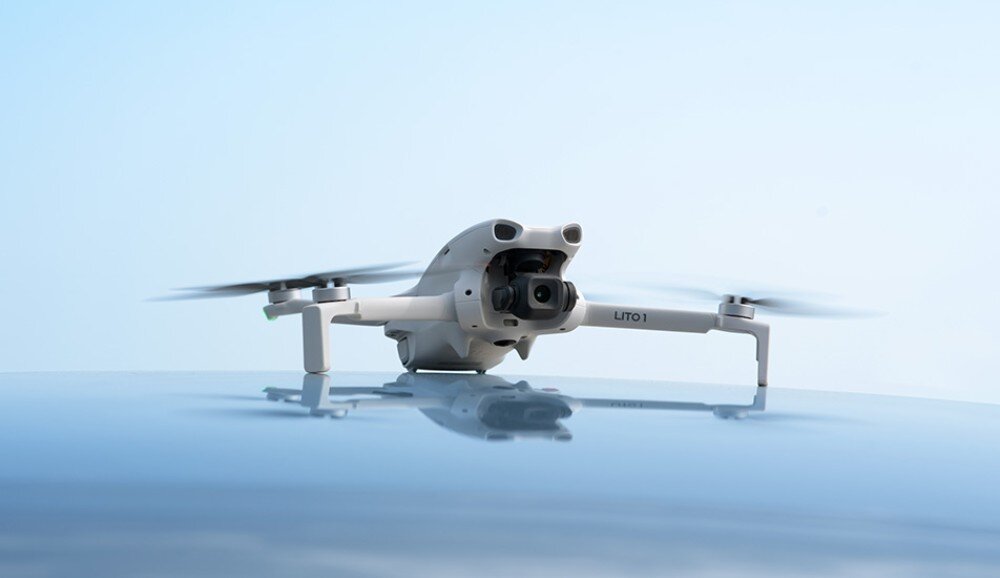 Dron DJI Lito 1 Szary dron stoi na gładkiej, odbijającej światło powierzchni pod bezchmurnym, jasnoniebieskim niebem. Kamera jest skierowana do przodu