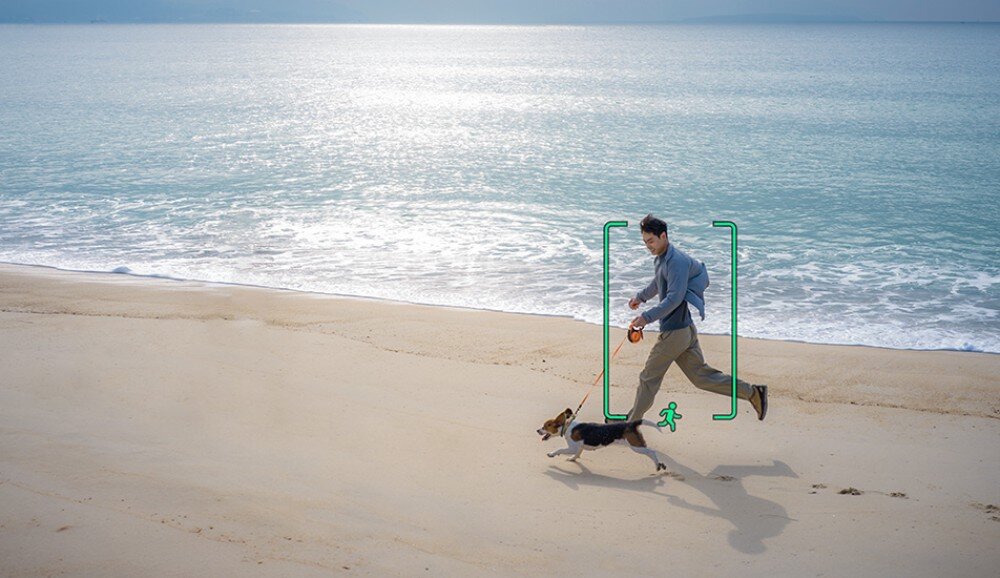 Dron DJI Lito 1 Widok z góry na piaszczystą plażę i jasnoniebieskie morze. Na plaży mężczyzna w szarej bluzie i beżowych spodniach biega z psem rasy beagle na smyczy. Sylwetki mężczyzny i psa są otoczone zielonym prostokątem