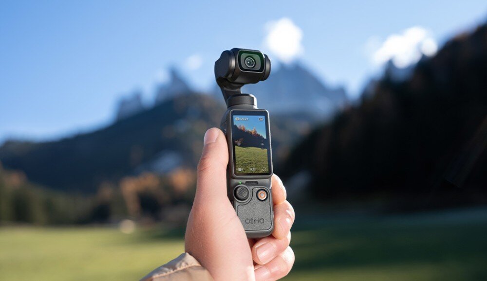 Kamera sportowa DJI OSMO POCKET 4 dłoń trzymająca urządzenie z włączonym ekranem, na którym widać góry, tło stanowi rozmyty krajobraz z górami