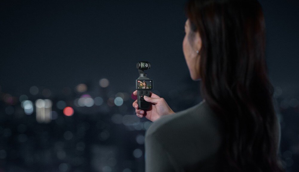 Kamera sportowa DJI OSMO POCKET 4 osoba widziana od tyłu trzymająca urządzenie z włączonym ekranem skierowane na rozmyte światła miasta w nocy