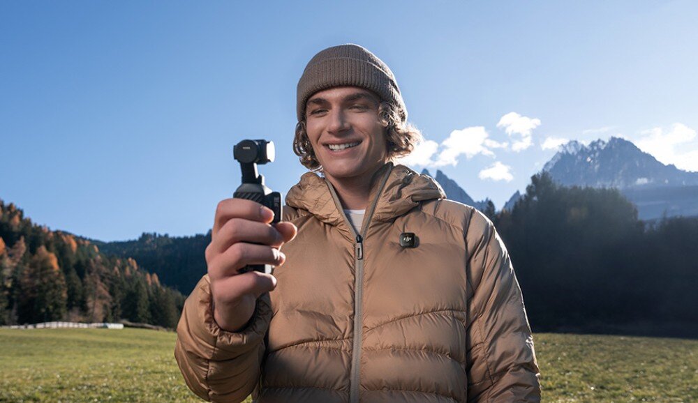 Kamera sportowa DJI OSMO POCKET 4 uśmiechnięty mężczyzna w czapce i kurtce trzymający urządzenie z kamerą, w tle widać góry i las