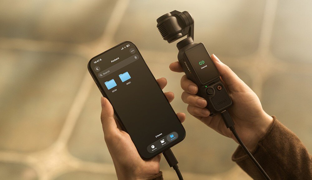 Kamera sportowa DJI OSMO POCKET 4 dwie dłonie trzymające smartfon z otwartym menedżerem plików i podłączone do niego kablem urządzenie z kamerą