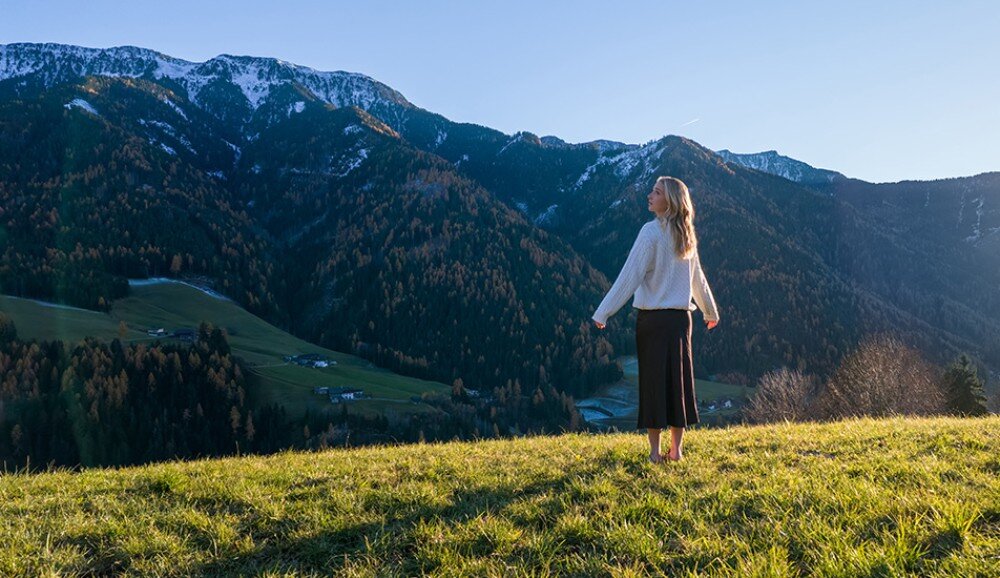 Kamera sportowa DJI OSMO POCKET 4 osoba stojąca na trawiastym zboczu, skierowana plecami do obiektywu, patrząca w górę, w tle widać wysokie góry