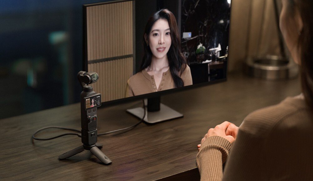Kamera sportowa DJI OSMO POCKET 4 osoba siedząca przy biurku przed monitorem, na którym widać twarz kobiety, obok monitora stoi urządzenie z kamerą na trójnogu