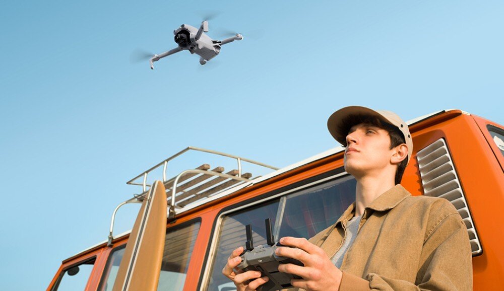 Dron DJI Lito X1 młody mężczyzna w czapce z daszkiem trzyma kontroler i patrzy w górę na lecącego drona obok pomarańczowego samochodu kempingowego