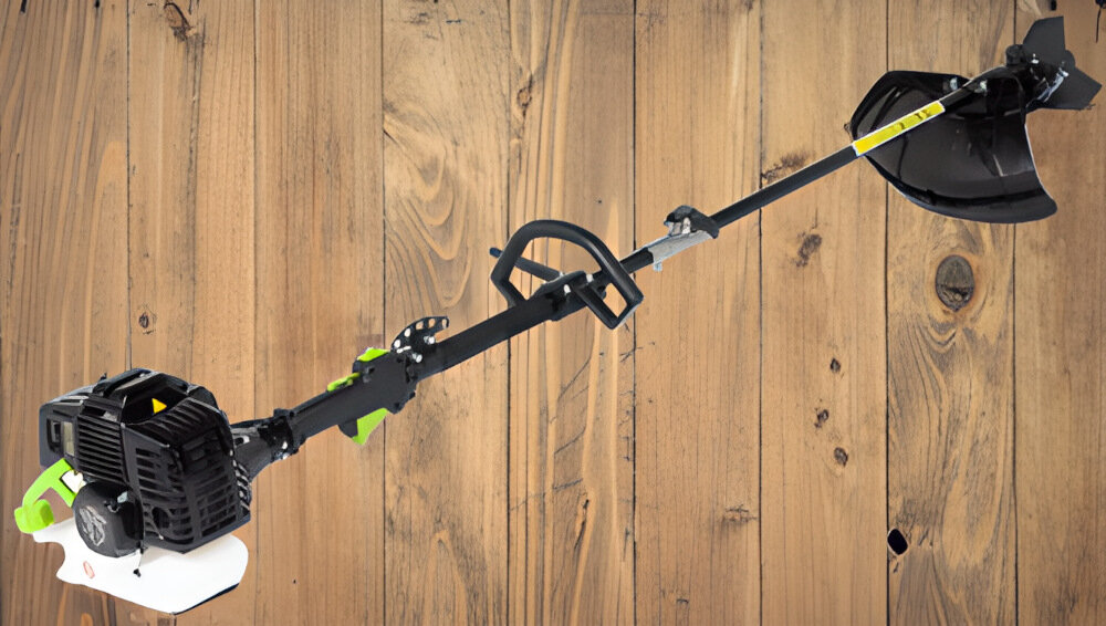 Kosa spalinowa z czarnym silnikiem i uchwytem, posiada osłonę tnącą oraz elementy w kolorze zielonym. Tło stanowi drewniana powierzchnia.