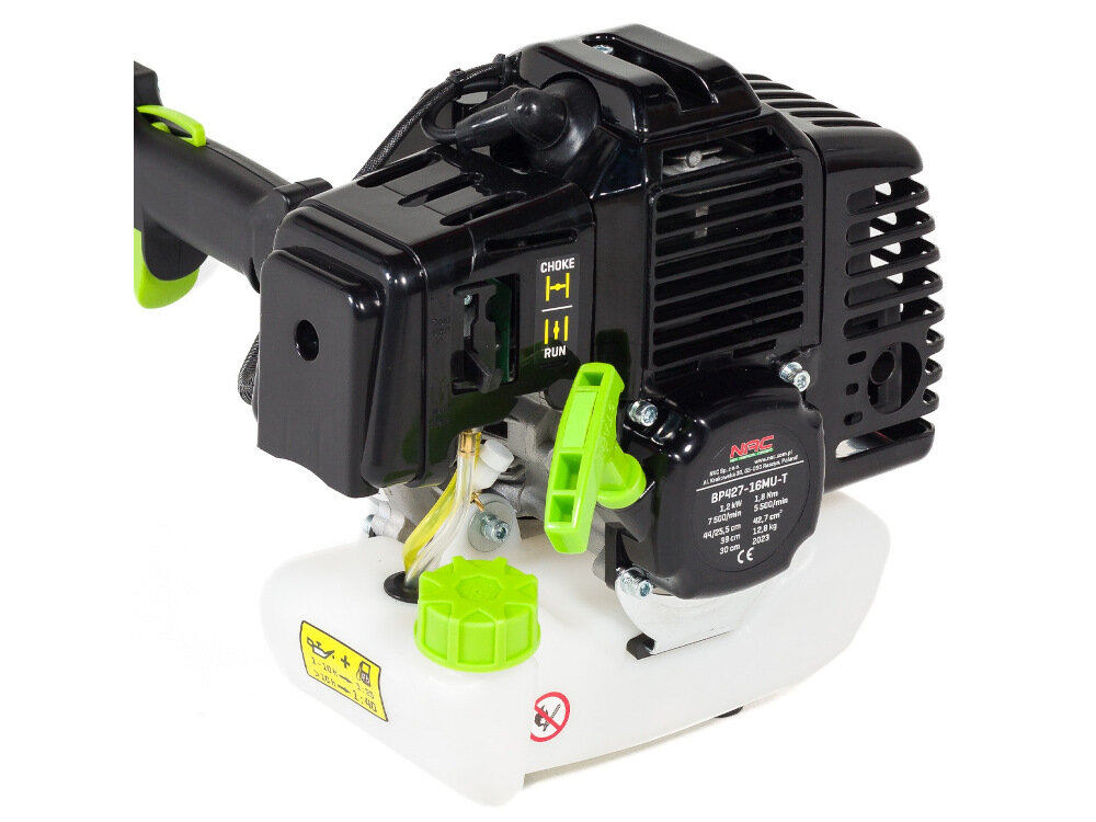 Czarna obudowa silnika z widocznymi napisami \'CHOKE\' i \'RUN\' oraz zielonymi elementami. Na białym zbiorniku znajduje się żółta naklejka z instrukcją i symbol zakazu palenia.