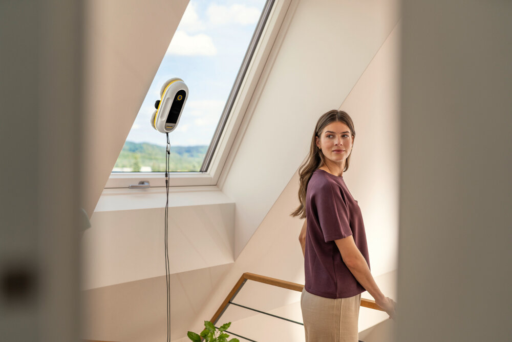 Na zdjęciu widać robota do mycia okien przymocowanego do dużego okna dachowego. W tle znajduje się osoba stojąca na schodach, patrząca w stronę robota.