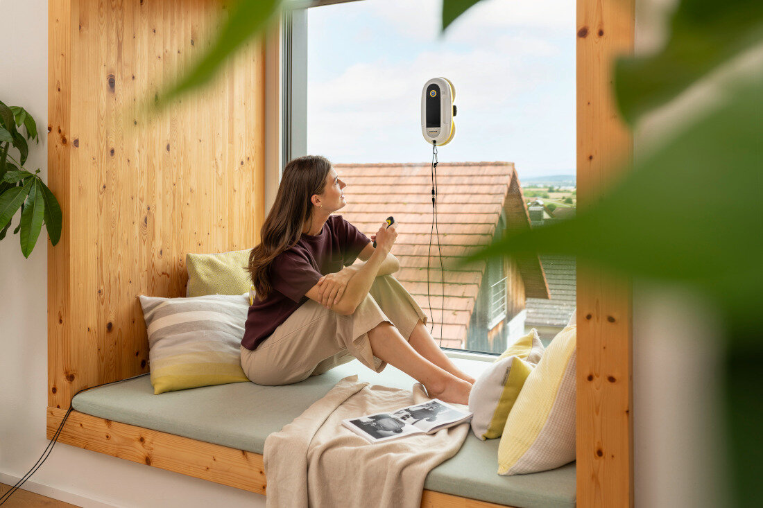 Kobieta siedzi na parapecie przy dużym oknie, patrząc na robota do mycia okien przymocowanego do szyby. W tle widać dachy budynków i krajobraz za oknem.