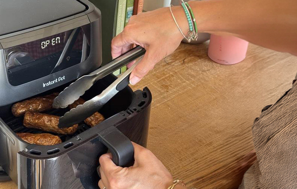 Osoba używa szczypiec do wyjmowania jedzenia z frytkownicy beztłuszczowej Instant Pot, na której wyświetlaczu widoczny jest napis \'OPEN\'.
