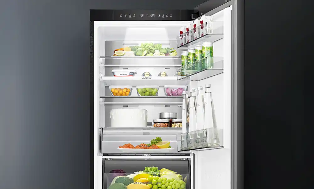 Otwarta lodówka z widocznymi półkami wypełnionymi różnorodnymi produktami spożywczymi, takimi jak owoce, warzywa, napoje i pojemniki z jedzeniem. Na drzwiach znajdują się butelki i kartony z napojami.