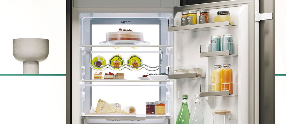 Otwarta lodówka z widocznymi półkami, na których znajdują się różne produkty spożywcze, takie jak napoje, ciasto i sery. Na górnej części lodówki widoczny jest napis \'ABT\'.