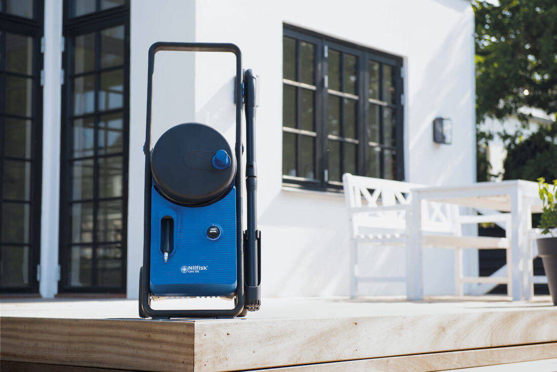 Na drewnianym tarasie stoi niebieska myjka ciśnieniowa z czarnymi elementami i logo \'NILFISK\'. W tle widoczny jest biały budynek z dużymi oknami i białą ławką.