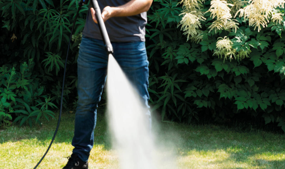Osoba używa myjki ciśnieniowej, z której wydobywa się strumień wody. W tle widoczne są zielone krzewy i trawnik.