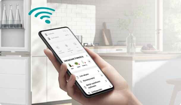 Osoba trzyma smartfon z aplikacją do zarządzania urządzeniami, na ekranie widoczny jest tekst \'Zarządzaj urządzeniami\'. W tle znajduje się nowoczesna kuchnia z białymi szafkami i elementami dekoracyjnymi.