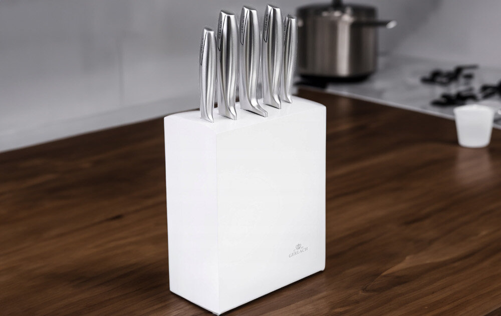 Na drewnianym blacie kuchennym stoi biały blok z pięcioma nożami o metalicznych uchwytach. Na bloku widoczne jest logo \'GERLACH\'.