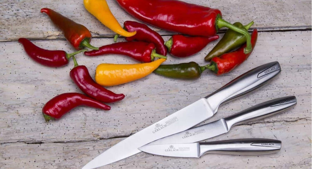 Na drewnianym tle leżą trzy noże z metalowymi uchwytami, obok nich różnokolorowe papryczki. Na ostrzach noży widoczne jest logo \'GERLACH\'.