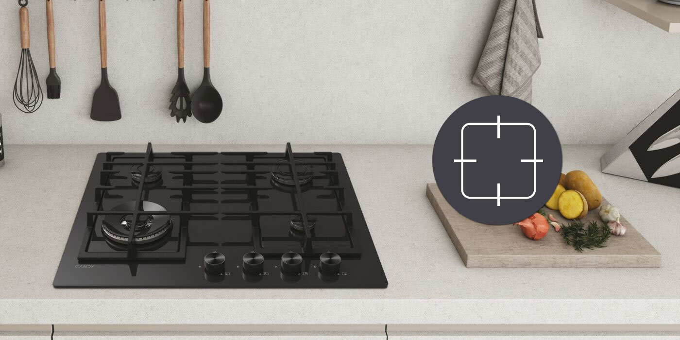 Na blacie kuchennym znajduje się czarna płyta gazowa z czterema palnikami i pokrętłami. Obok leży deska do krojenia z warzywami, a nad blatem wiszą kuchenne akcesoria.