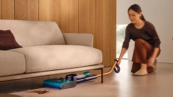 Kobieta trzyma w ręku mop, który jest złożony równolegle do podłogi i czyści przestrzeń pod sofą