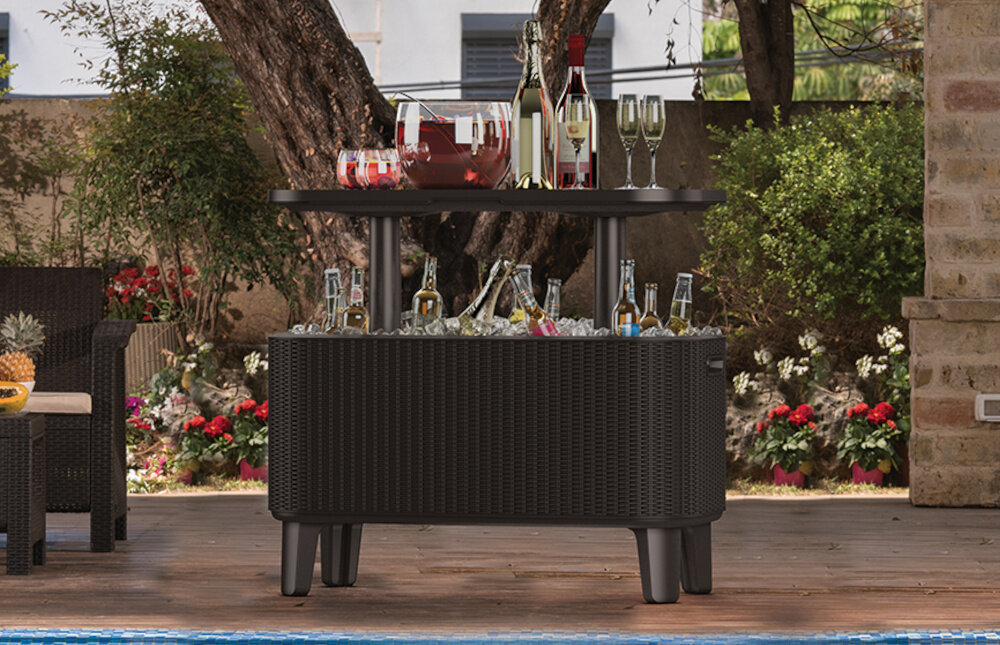 Na drewnianym tarasie stoi antracytowy stolik z plecionki, na którym znajdują się butelki i kieliszki. W tle widoczne są drzewa i kwiaty w ogrodzie.