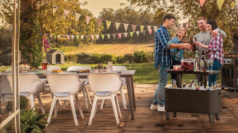 Na drewnianym tarasie znajduje się zestaw mebli ogrodowych z białymi krzesłami i stołem, a obok stoi stolik z napojami i lodem. W tle widać kolorowe girlandy i zielony krajobraz.