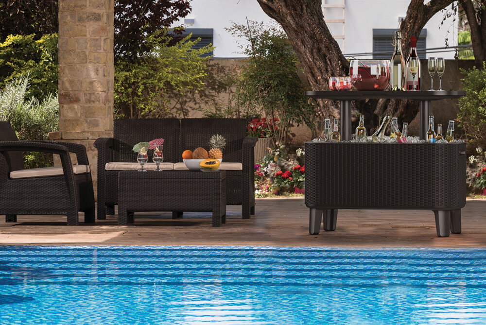 Na tarasie przy basenie znajduje się zestaw mebli ogrodowych z ciemnego rattanu, w tym stolik z napojami i przekąskami. W tle widoczne są drzewa i krzewy ozdobne.