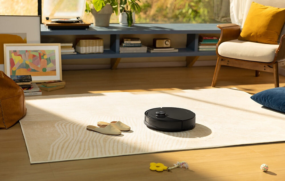 Czarny robot sprzątający znajduje się na jasnym dywanie w salonie. W tle widać półkę z książkami i dekoracjami oraz fotel z poduszką.