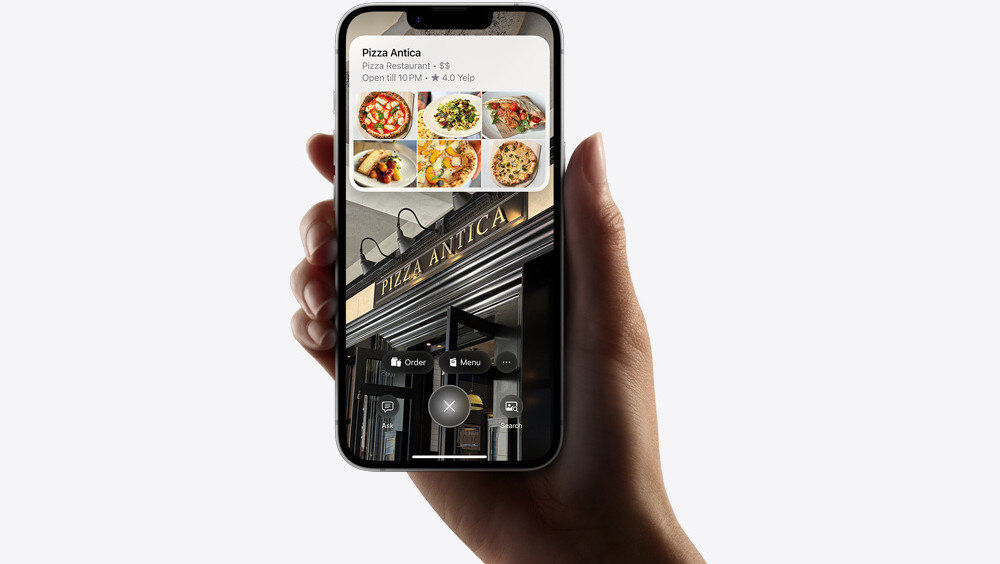 Osoba trzyma smartfon z wyświetlonym na ekranie interfejsem aplikacji restauracyjnej. Na ekranie widoczny jest tekst: \'Pizza Antica, Pizza Restaurant, $$, Open til 10PM, 4.0 Yelp\'.