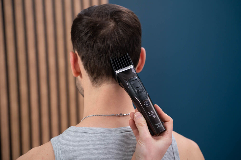 Osoba używa strzyżarki do przycinania włosów na karku innej osoby. Strzyżarka jest czarna z widocznymi przyciskami i logo producenta GÖTZE & JENSEN.