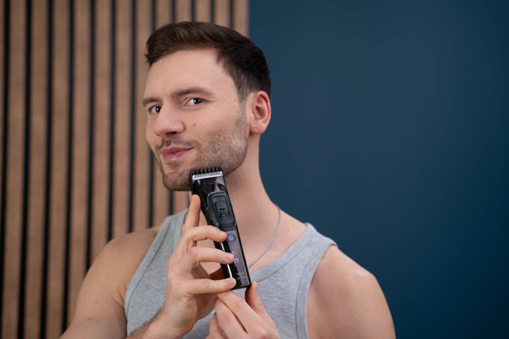Mężczyzna trzyma strzyżarkę przy twarzy, prezentując jej użycie. W tle widoczna jest drewniana ściana i niebieska powierzchnia.