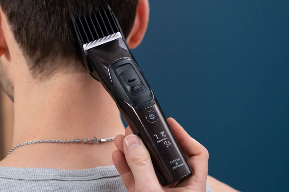 Osoba używa strzyżarki do włosów, trzymając ją przy karku. Na urządzeniu widoczne są przyciski oraz napis \'Götze & Jensen\'.