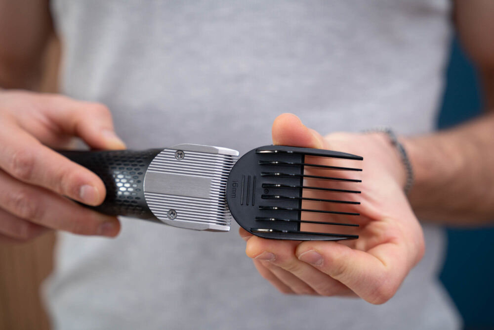 Osoba trzyma strzyżarkę z czarną rączką i metalową głowicą, do której przymocowany jest czarny grzebień z oznaczeniem \'16 mm\'.