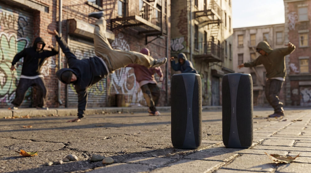 Na ulicy widać dwóch czarnych głośników przenośnych z logo XMUSIC. W tle kilku ludzi wykonuje taneczne ruchy na tle budynków pokrytych graffiti.
